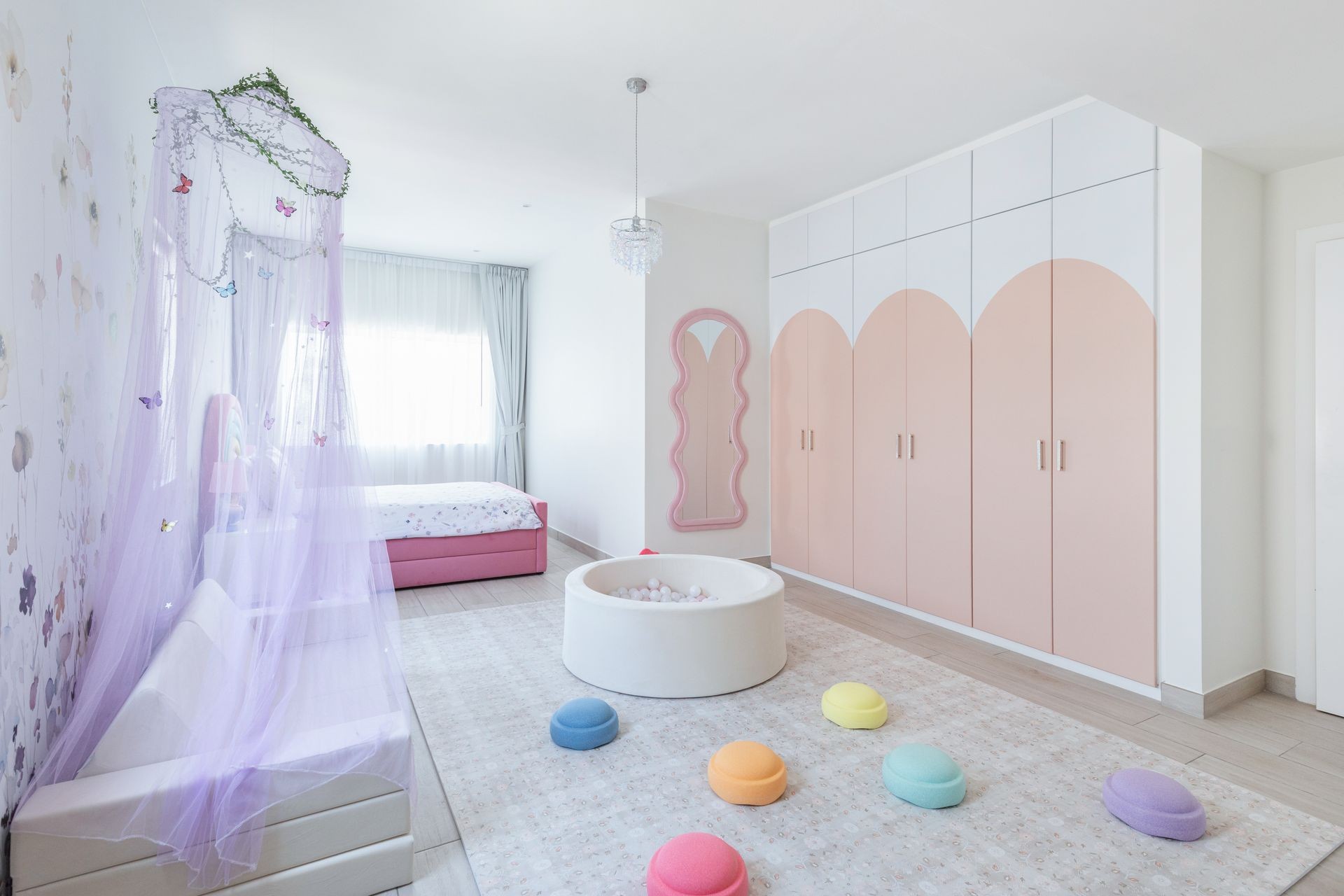 Children’s bedroom in pastel tones with a butterfly canopy, rainbow wardrobes, soft seating, and playful textures.