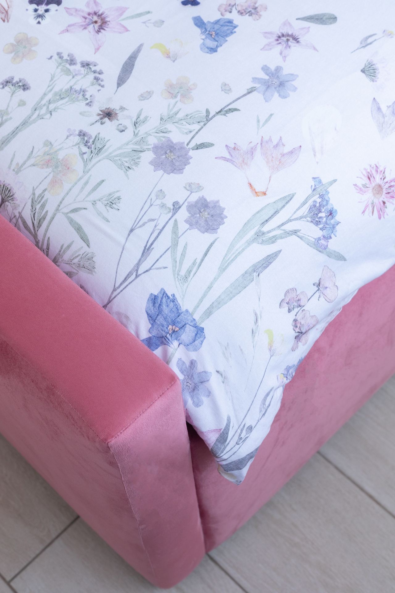 Floral-patterned bedsheet on a pink velvet bed corner, on a light wooden floor.
